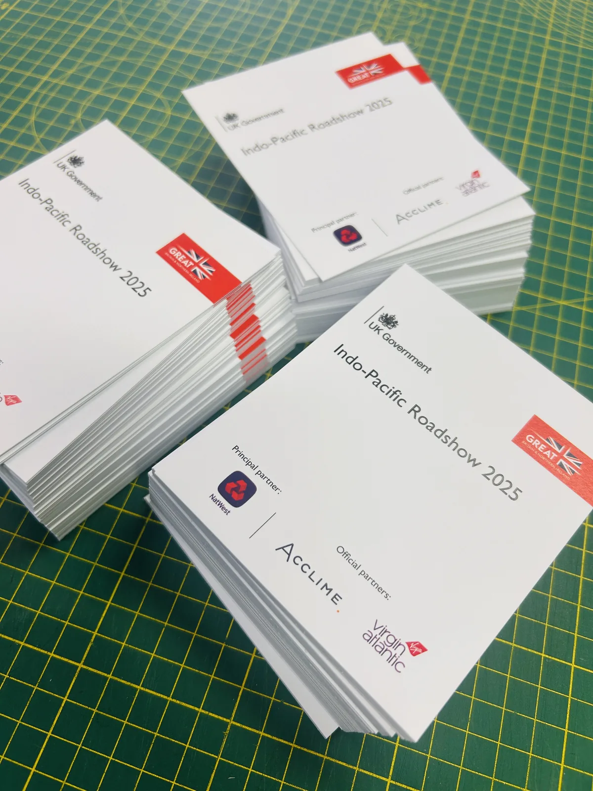 Three stacks of printed delegate badges for a UK Government Indo-Pacific Roadshow, on a green cutting mat