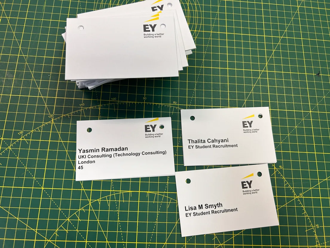 Row of white EY-branded delegate badges laid out on a cutting mat, each with a different name and job title