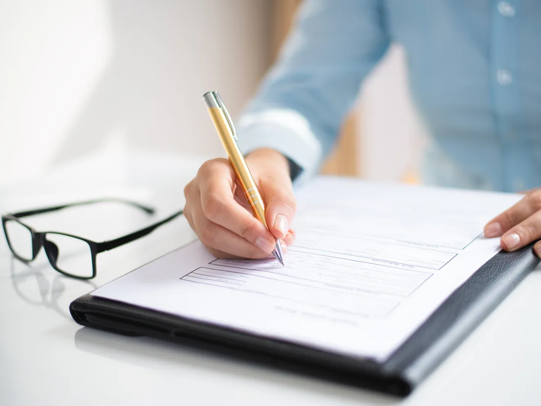 Person writing on a document held in a folder, reading glasses beside it