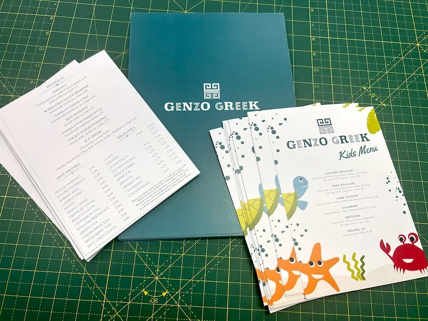 Greek restaurant menus with seafood illustrations, laid out on a green cutting mat