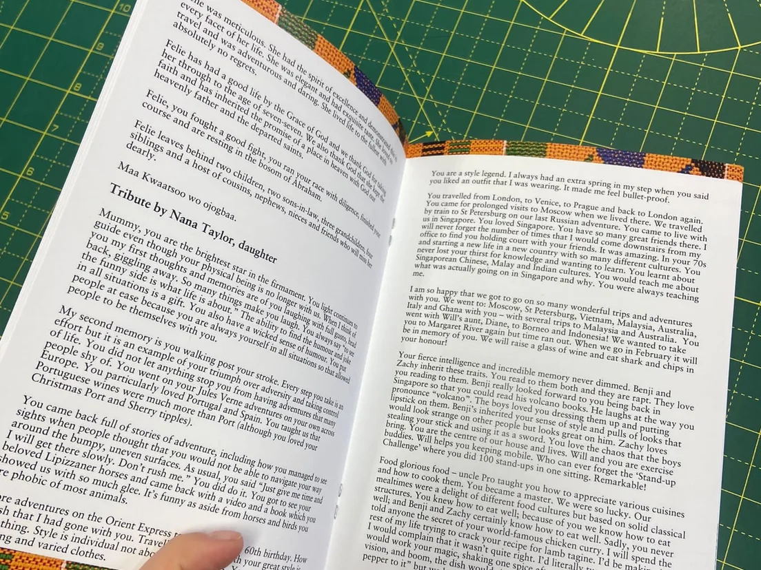 Hands holding an order of service booklet open at a printed text spread, on a green cutting mat