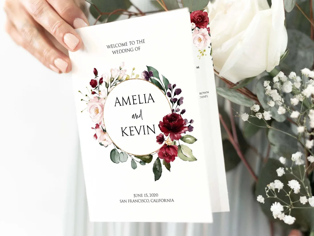 Order of service with a burgundy floral cover, held in two hands beside white roses