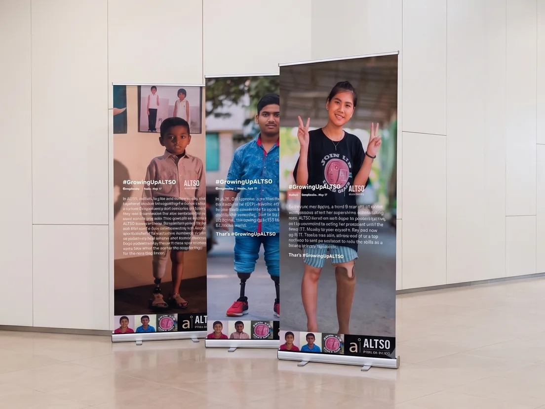 Three charity campaign roller banners standing in a row in an office foyer, each with a portrait and story text